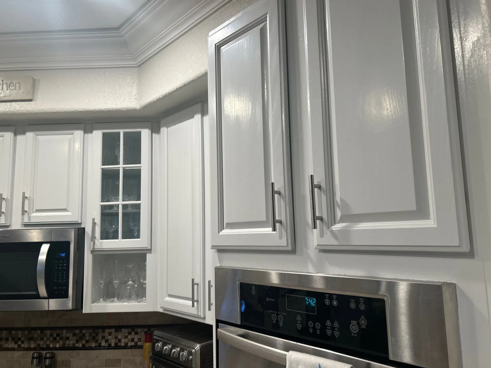 Cabinet Refinishing in Aberdeen, Florida. White painted raised-panel upper cabinets with crown moulding and a glass-front cabinet, beside stainless steel microwave and oven.