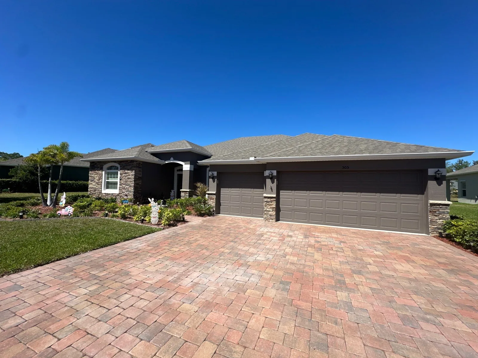 Exterior Painting in Bayside Lakes, Florida. Single-storey taupe stucco home with stone veneer accent, a three-bay garage and a brick paver driveway.