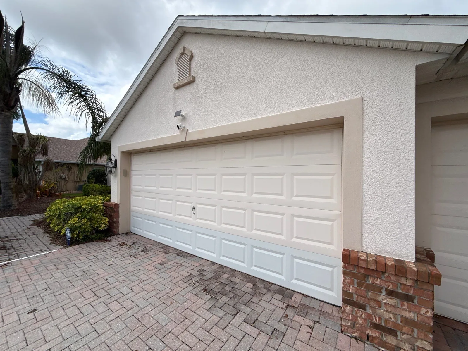 Exterior Painting in Bayside Lakes, Florida. Two-car garage with cream stucco walls, white sectional garage door and brick veneer column over a paver driveway.
