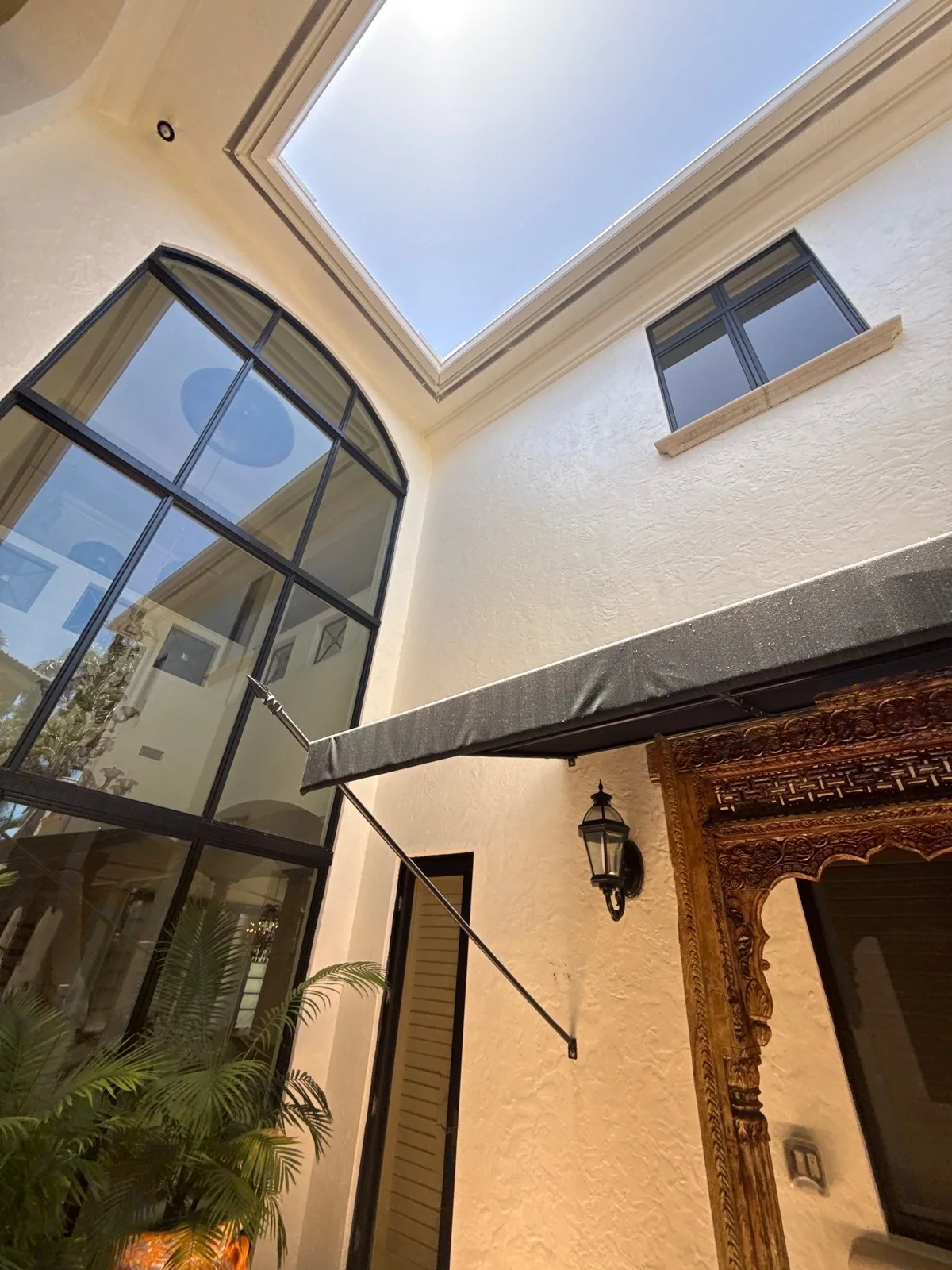 Exterior Painting in Boca Raton, Florida. Courtyard with white stucco walls, a tall black-framed arched window, black awning and a carved wood door surround.