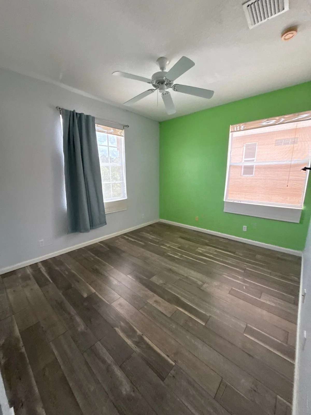 Interior Painting in Coral Gardens, Florida. Empty bedroom with a bright green accent wall, light grey walls, white ceiling with fan and grey wood-look flooring.