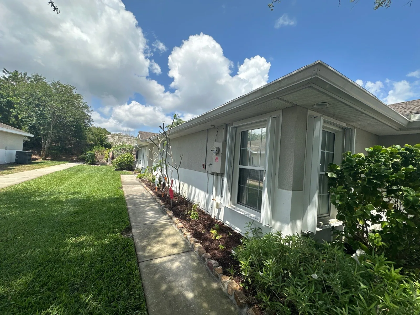 Exterior Painting in Eau Gallie, Florida. Side elevation of a single-storey home with light stucco walls, white trim and gutters, walkway and lawn under a blue sky.