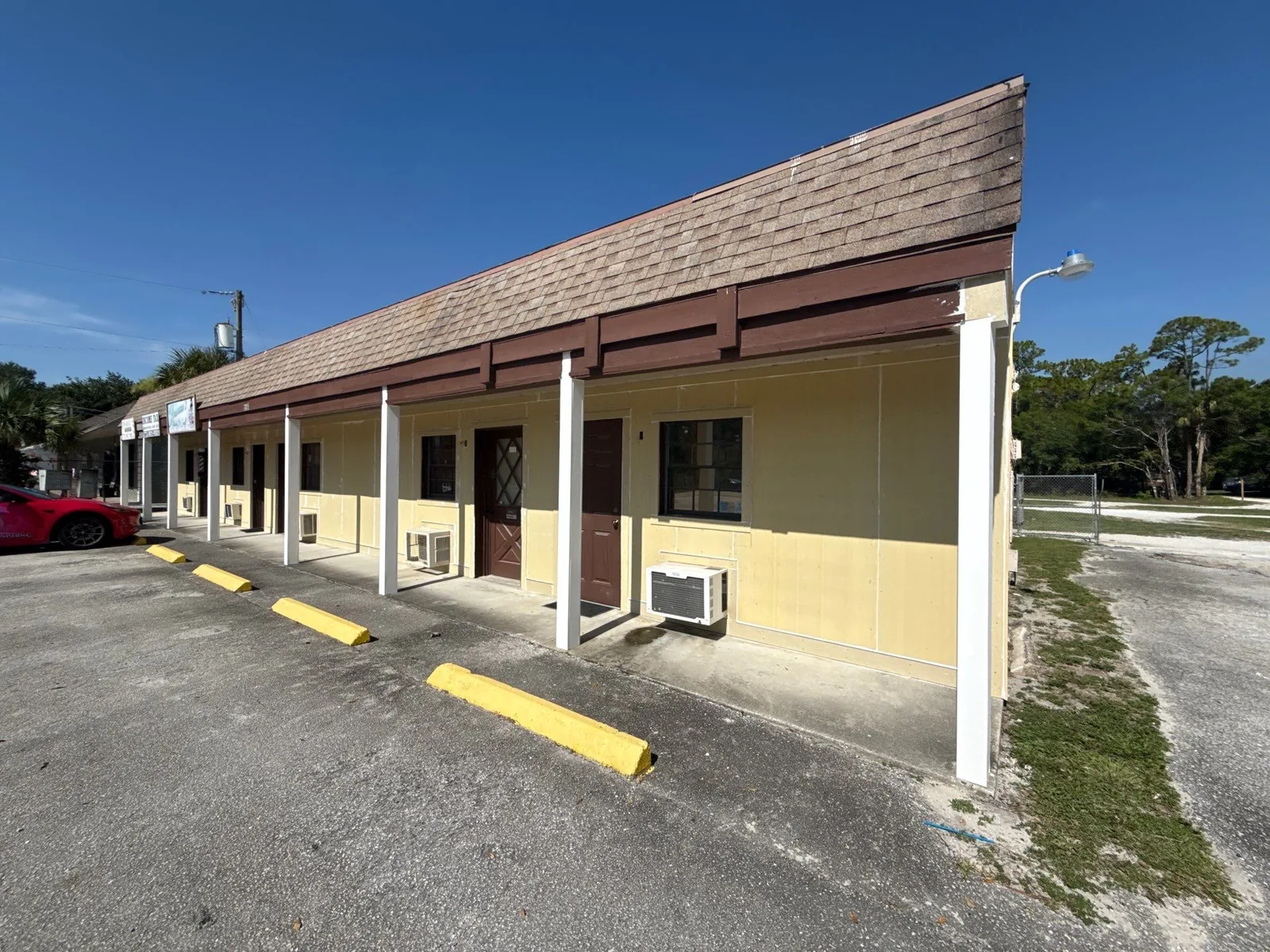 Commercial Painting in Florida Ridge, Florida. Single-storey commercial strip building painted cream and pale yellow with brown trim and white columns under a shingle roof, beside a paved parking area.
