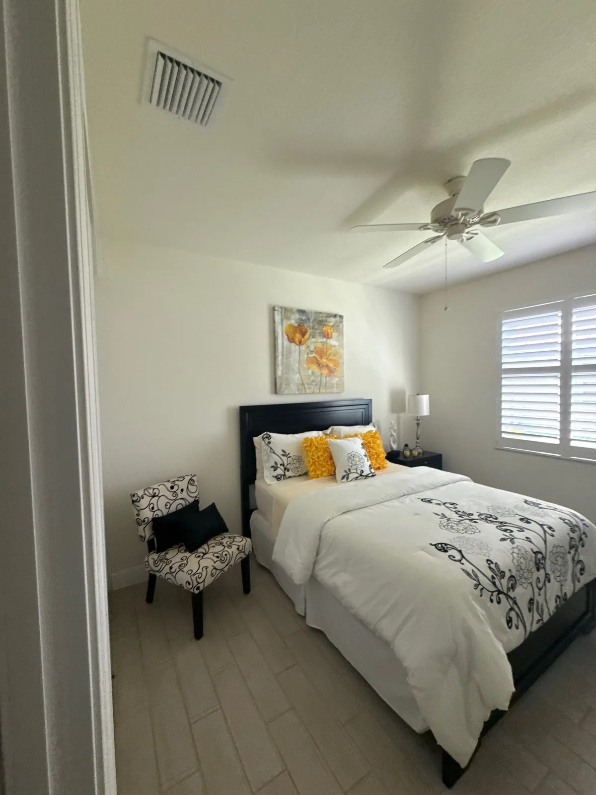 Interior Painting in Florida Ridge, Florida. Staged bedroom viewed from the doorway, painted walls and ceiling, made bed, accent chair, ceiling fan and shuttered window.