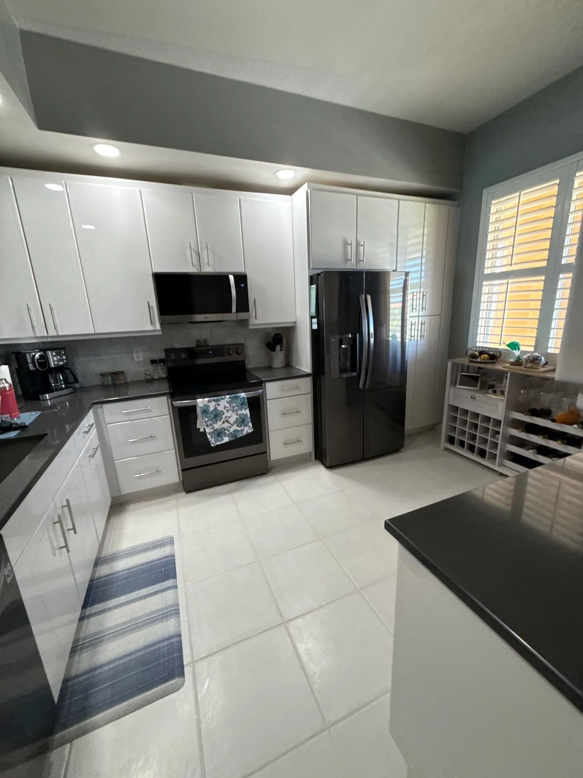 Cabinet Refinishing in Fort Pierce, Florida. Kitchen with white painted cabinets, dark countertops, stainless steel refrigerator and range, plantation shutters and large-format floor tile.