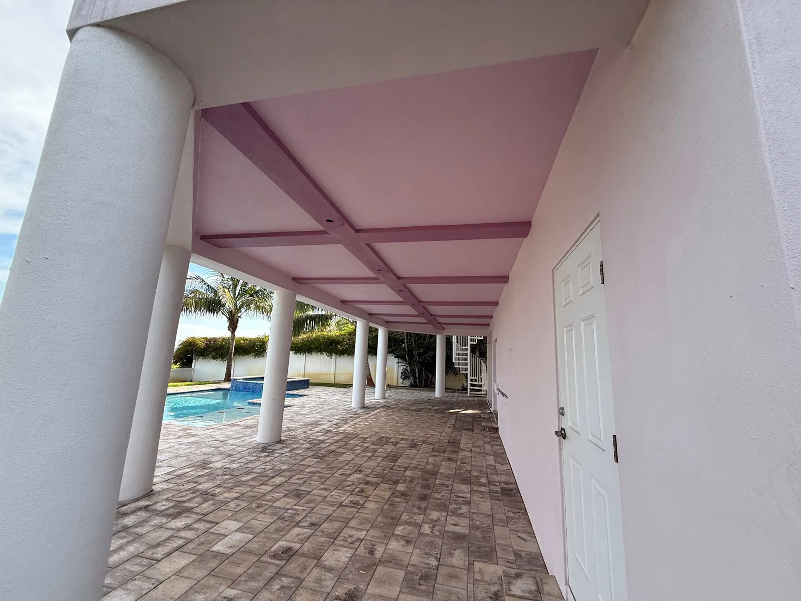 Exterior Painting in Fort Pierce, Florida. Covered ground-level breezeway with pink painted ceiling and beams, white columns, paver floor and pool beyond.