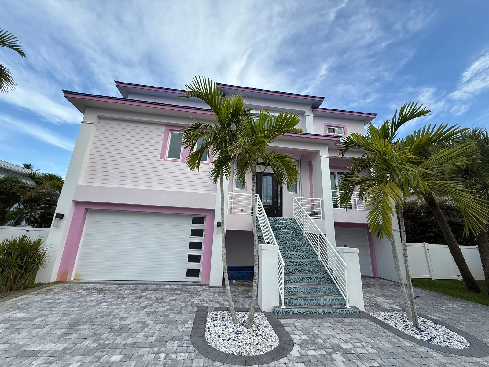 Exterior Painting in Fort Pierce, Florida. Two-storey house painted white with pink trim, white garage doors, a tiled exterior staircase with white railings, paver driveway and palms.