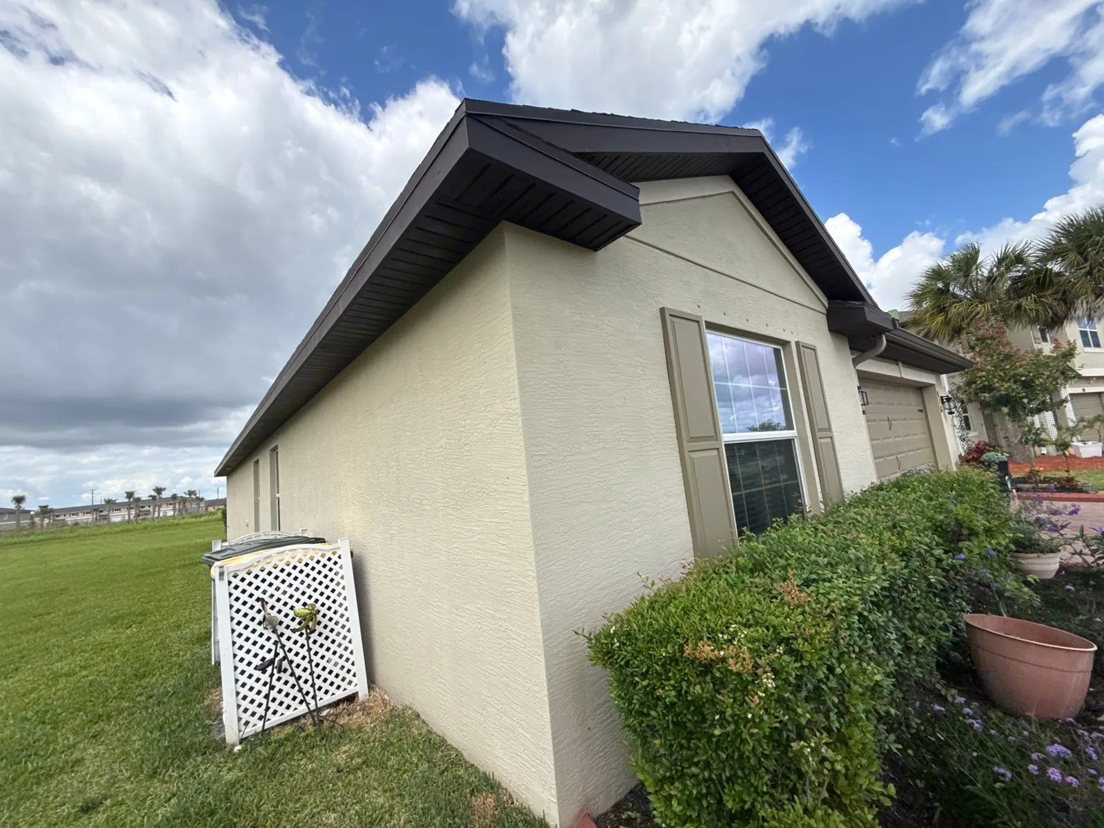 Exterior Painting in Fort Pierce, Florida. Corner of a single-storey stucco house painted cream with dark brown fascia and window shutters, lawn and shrubs in front.