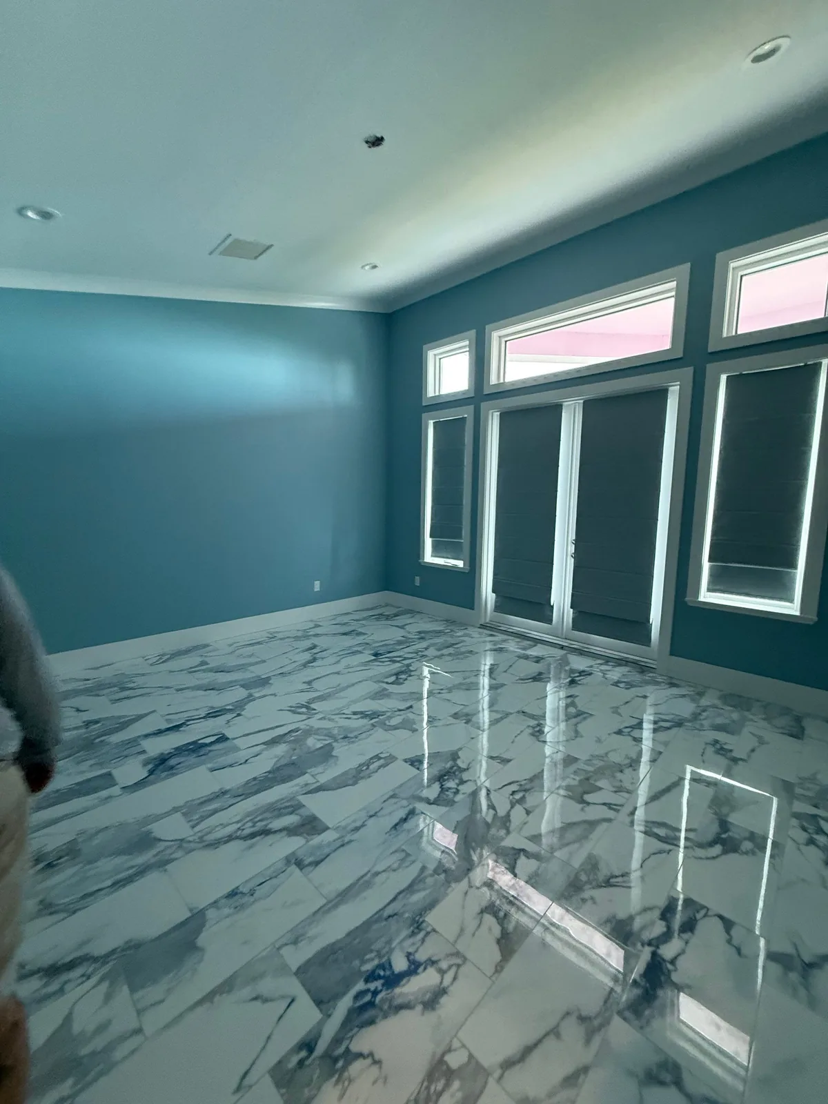 Interior Painting in Fort Pierce, Florida. Large empty room with teal walls, white crown moulding, tall shaded windows and polished marble-look tile floor.