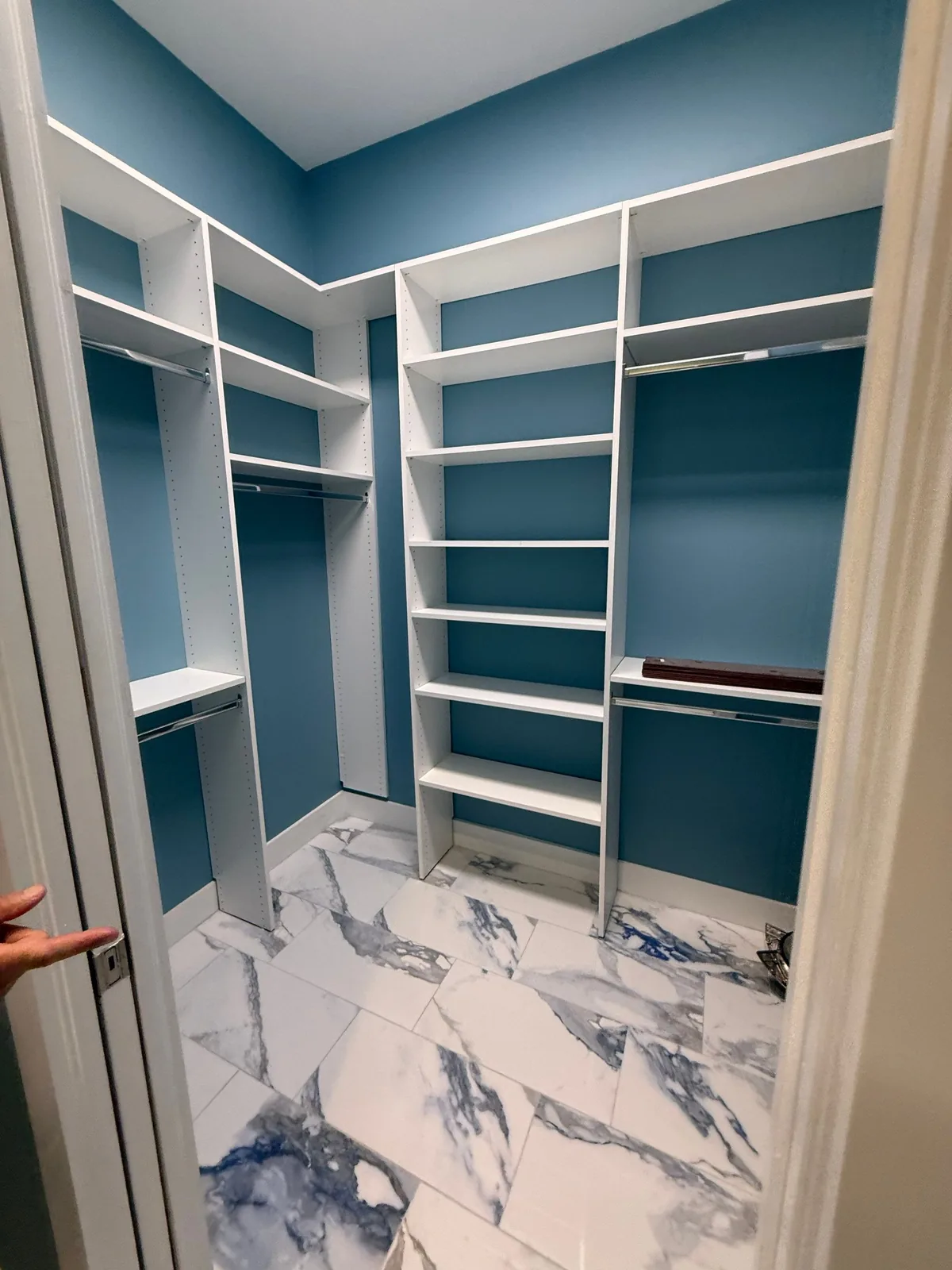 Interior Painting in Fort Pierce, Florida. Walk-in closet with blue walls, white built-in shelving and hanging rods, marble-look tile floor.