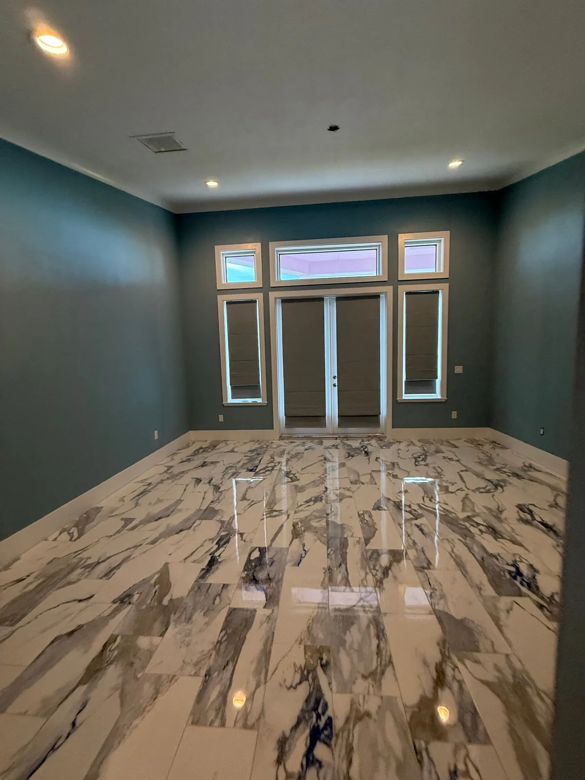 Interior Painting in Fort Pierce, Florida. Empty room with teal-painted walls, white crown moulding and trim, French doors with transom windows above, and a polished marble-look tile floor.