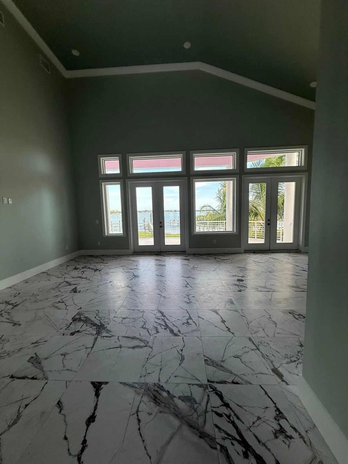 Interior Painting in Fort Pierce, Florida. Empty living room with dark teal walls, vaulted ceiling, a wall of windows and French doors, and a marble-look tile floor.