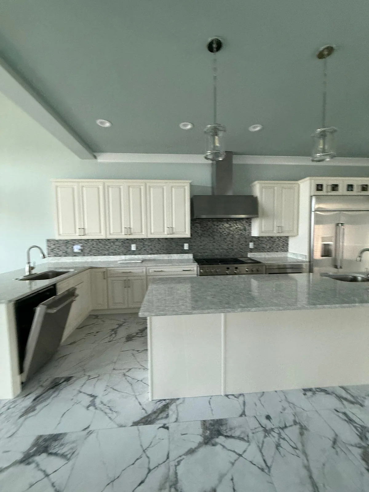 Interior Painting in Fort Pierce, Florida. Empty kitchen with pale sage-grey walls and ceiling, cream raised-panel cabinets, light stone countertops, mosaic tile backsplash and marble-look tile floor.
