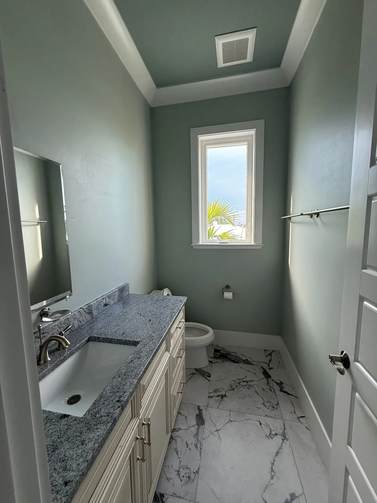 Interior Painting in Fort Pierce, Florida. Powder room with sage-green walls, white crown moulding and window trim, white vanity with grey stone countertop and marble-look tile floor.