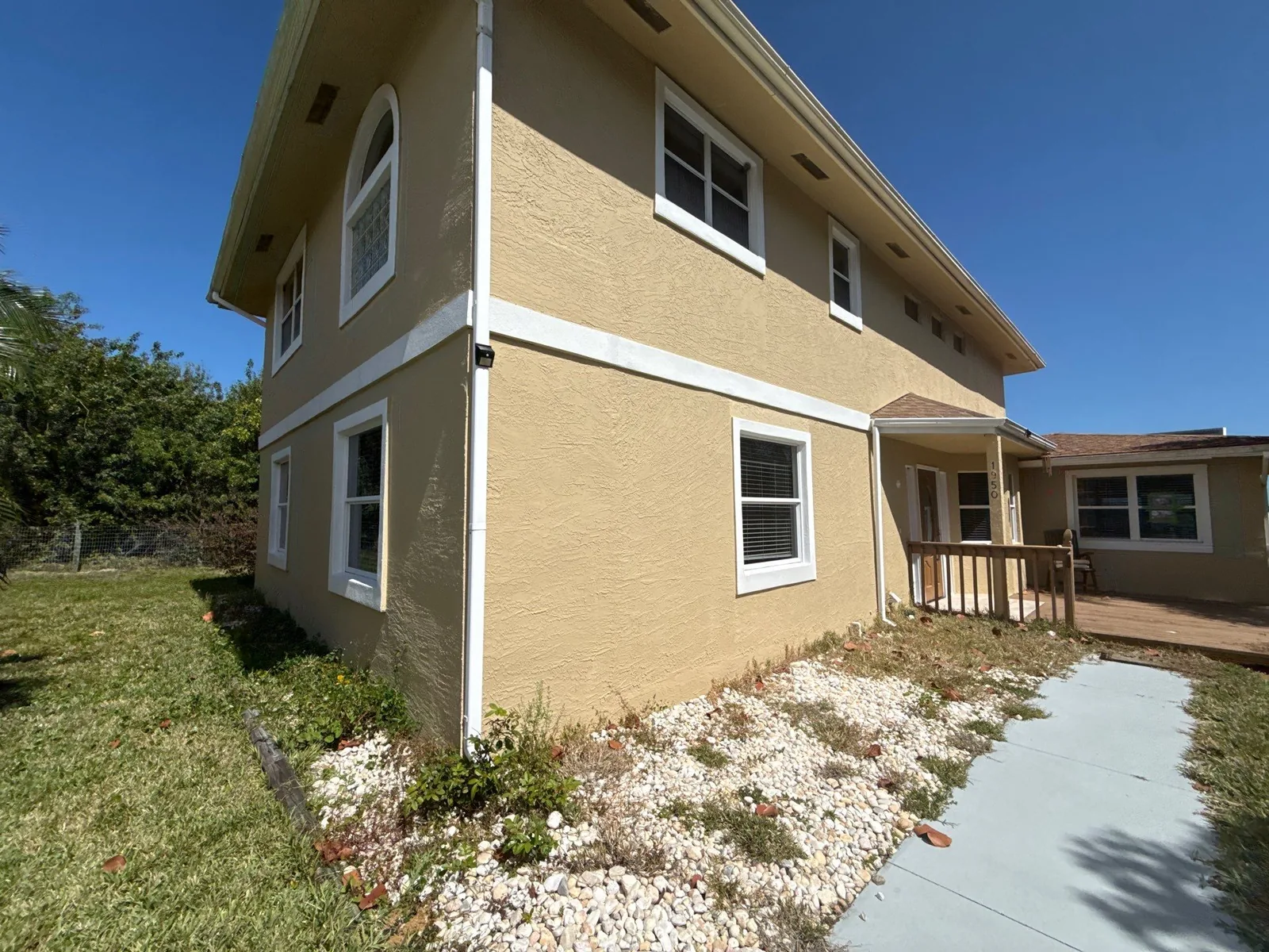 Exterior Painting in Gifford, Florida. Two-storey stucco house exterior painted tan with white trim and white downspouts, under a clear blue sky.