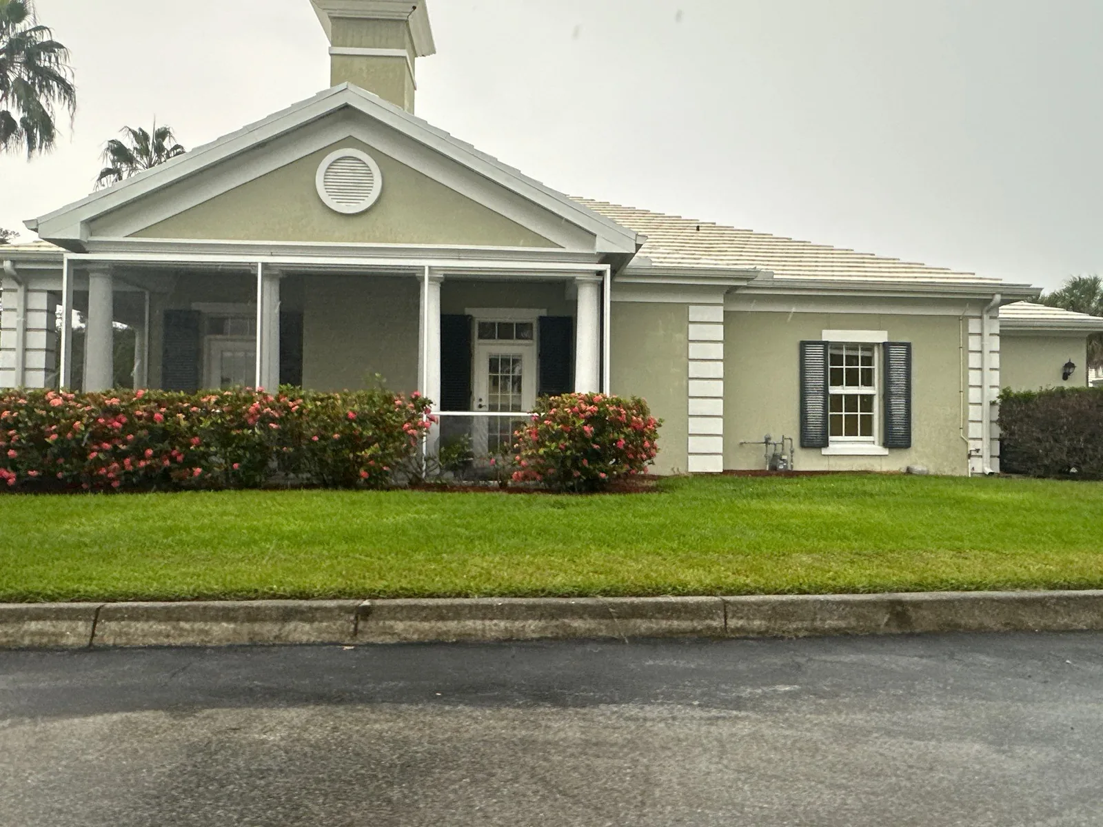 Exterior Painting in Gifford, Florida. Single-storey home with pale green stucco walls, white trim and columns, and a white portico entry above a flowering hedge.