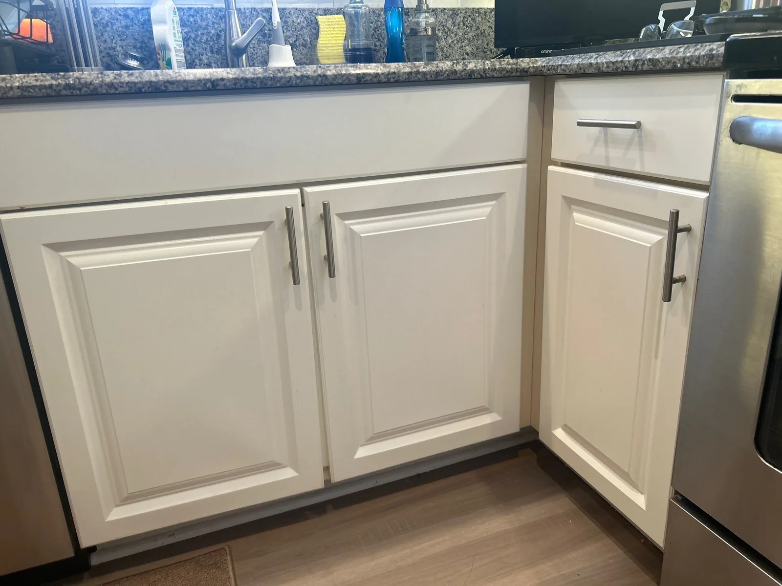 Cabinet Refinishing in Golden Lakes, Florida. Refinished white kitchen base cabinets with raised-panel doors, dark bar-pull hardware, granite countertop and wood-look flooring.
