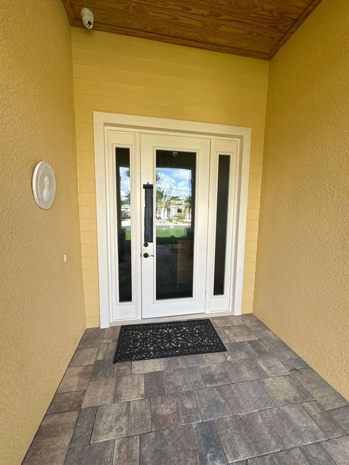 Exterior Painting in Gomez, Florida. Covered front entry with yellow stucco walls, white double glass entry doors with sidelights and white trim, wood-plank porch ceiling and a paver walkway.