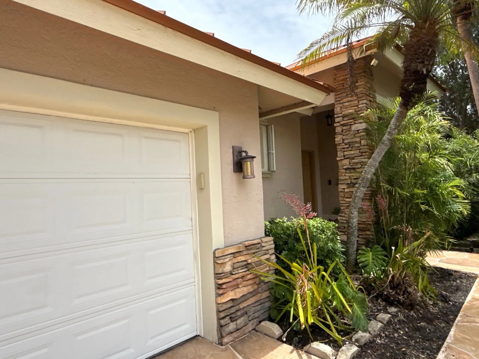 Exterior Painting in Gomez, Florida. Single-storey home exterior with beige stucco, white garage door, stone veneer base and a wall lantern beside the entry, with palms and landscaping.