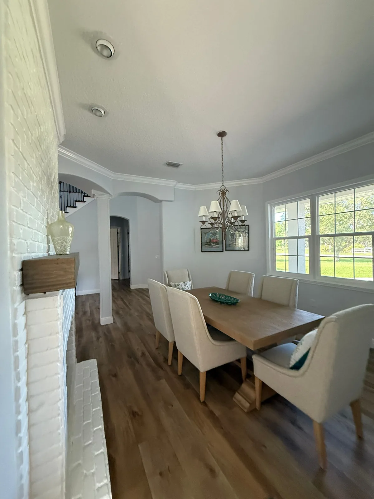 Interior Painting in Grant-Valkaria, Florida. Furnished dining room with pale grey painted walls, white ceiling and crown moulding, white painted brick fireplace, wood table with upholstered chairs and a chandelier.