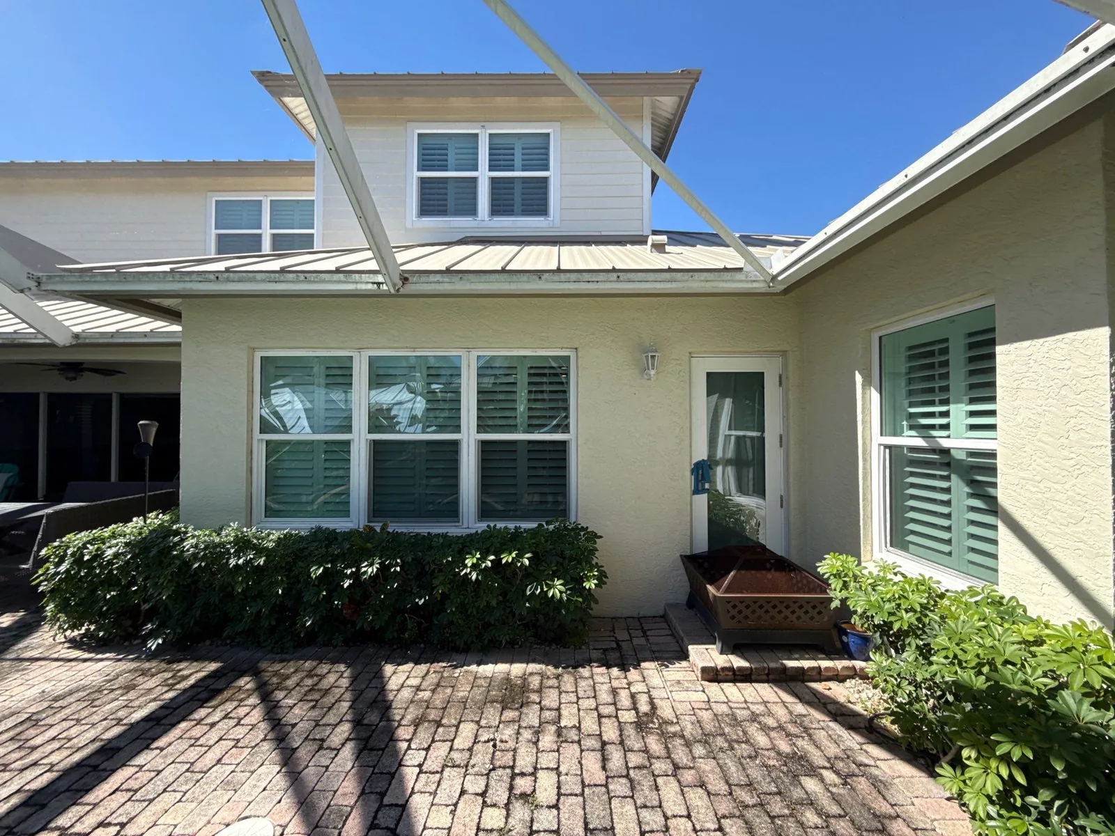 Exterior Painting in Hobe Sound, Florida. Rear of a home with pale yellow stucco walls, white window trim, green louvered shutters and a metal roof above a paver patio.