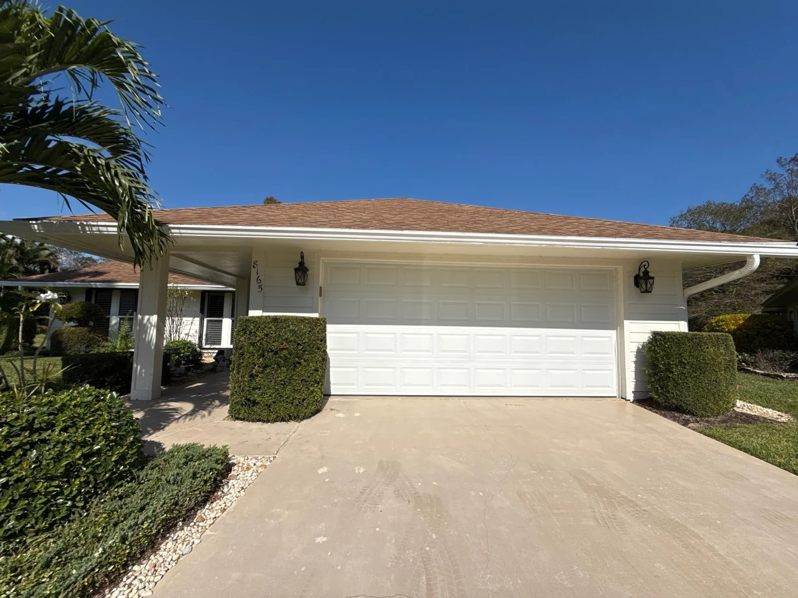 Exterior Painting in Hobe Sound, Florida. Single-storey home with freshly painted cream stucco exterior and white garage door, trimmed hedges and concrete driveway under a clear blue sky.