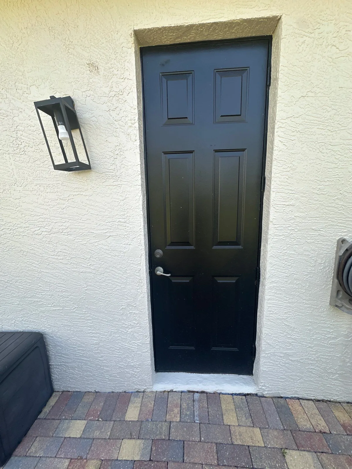 Exterior Painting in Indian River Estates, Florida. Black six-panel exterior door set in a white stucco wall, with a wall sconce and paver walkway.