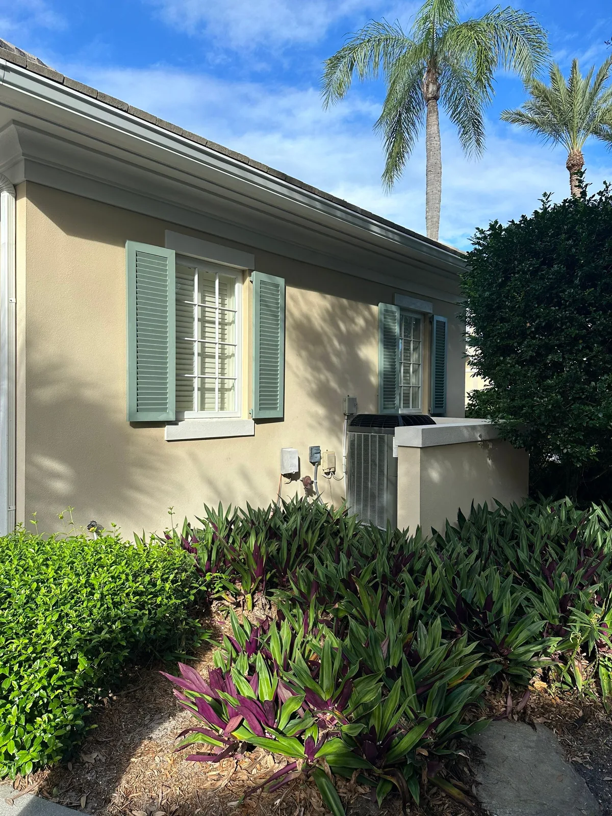 Exterior Painting in Indian River Shores, Florida. Single-storey stucco house exterior in cream with sage green window shutters and white trim, landscaping in front.