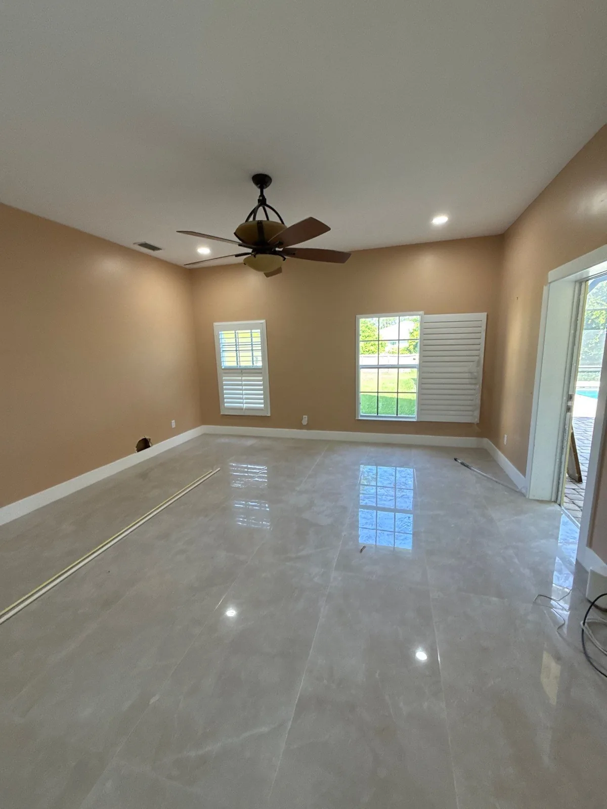 Interior Painting in Indrio, Florida. Empty living room with tan walls, white ceiling and baseboards, ceiling fan, two shuttered windows and polished tile floor.