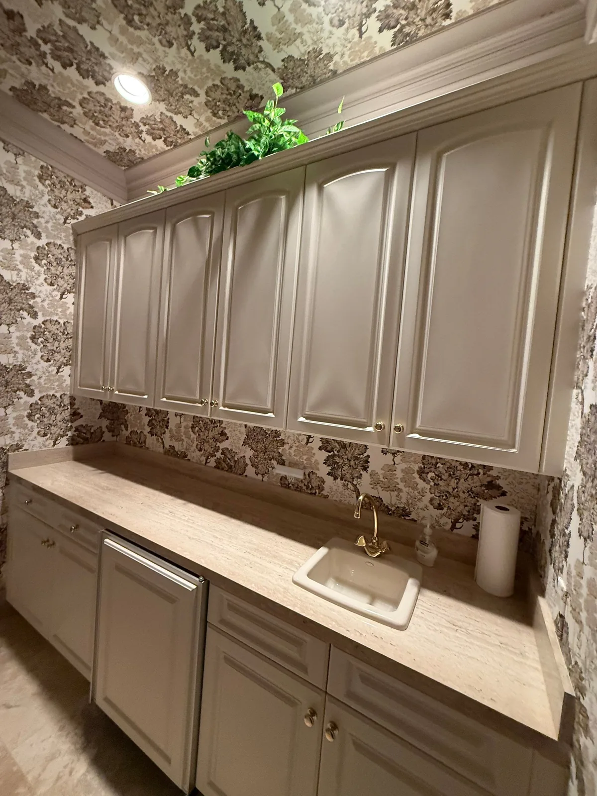 Cabinet Refinishing in Jensen Beach, Florida. Butler's pantry with cream raised-panel upper and lower cabinets, brass hardware, stone countertop, bar sink and patterned wallpaper.