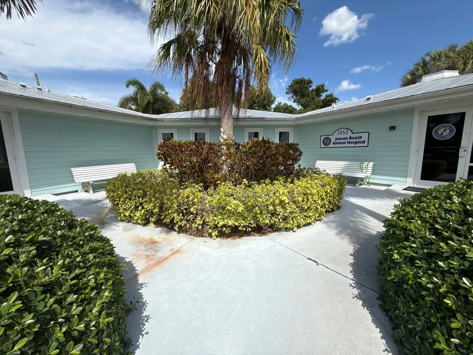 Commercial Painting in Jensen Beach, Florida. Courtyard of a pale green commercial building with white trim, white benches, palm tree and hedges on a concrete walkway.