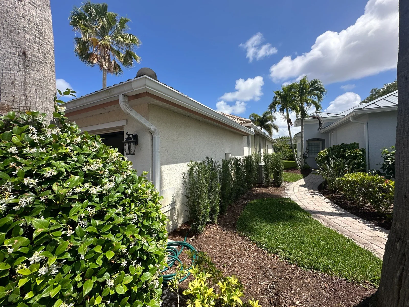 Exterior Painting in Jensen Beach, Florida. Side elevation of a single-storey stucco home in light cream with a barrel-tile roof, hedge row and paver walkway.