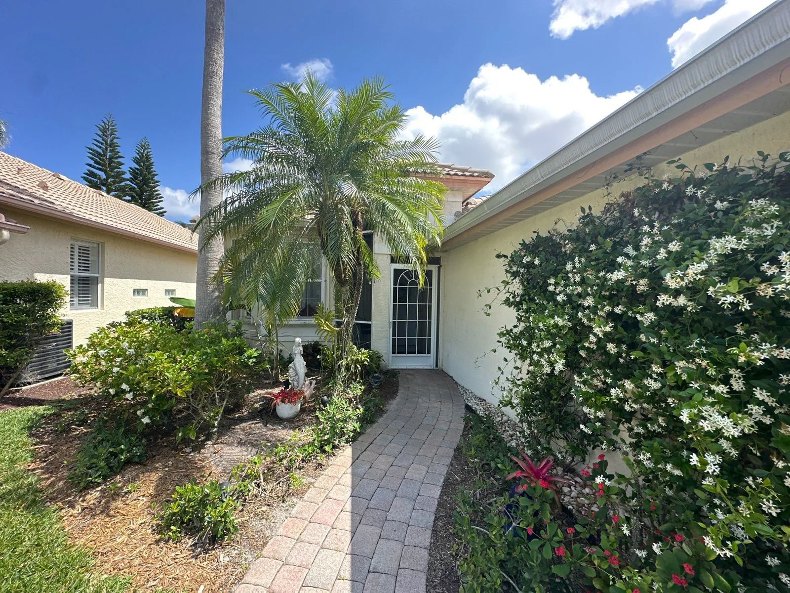 Exterior Painting in Jensen Beach, Florida. Front entry of a pale yellow stucco home with white door and trim, paver walkway and flowering shrubs.