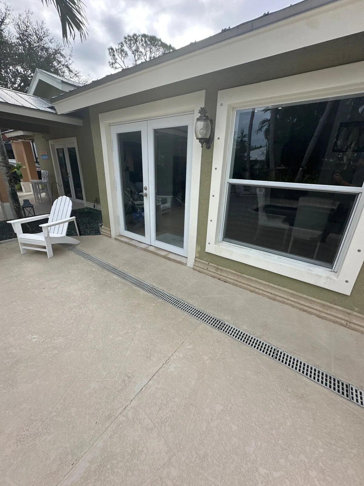 Exterior Painting in Jensen Beach, Florida. Exterior wall in olive green with white window and French door trim, beside a concrete patio with a white Adirondack chair.