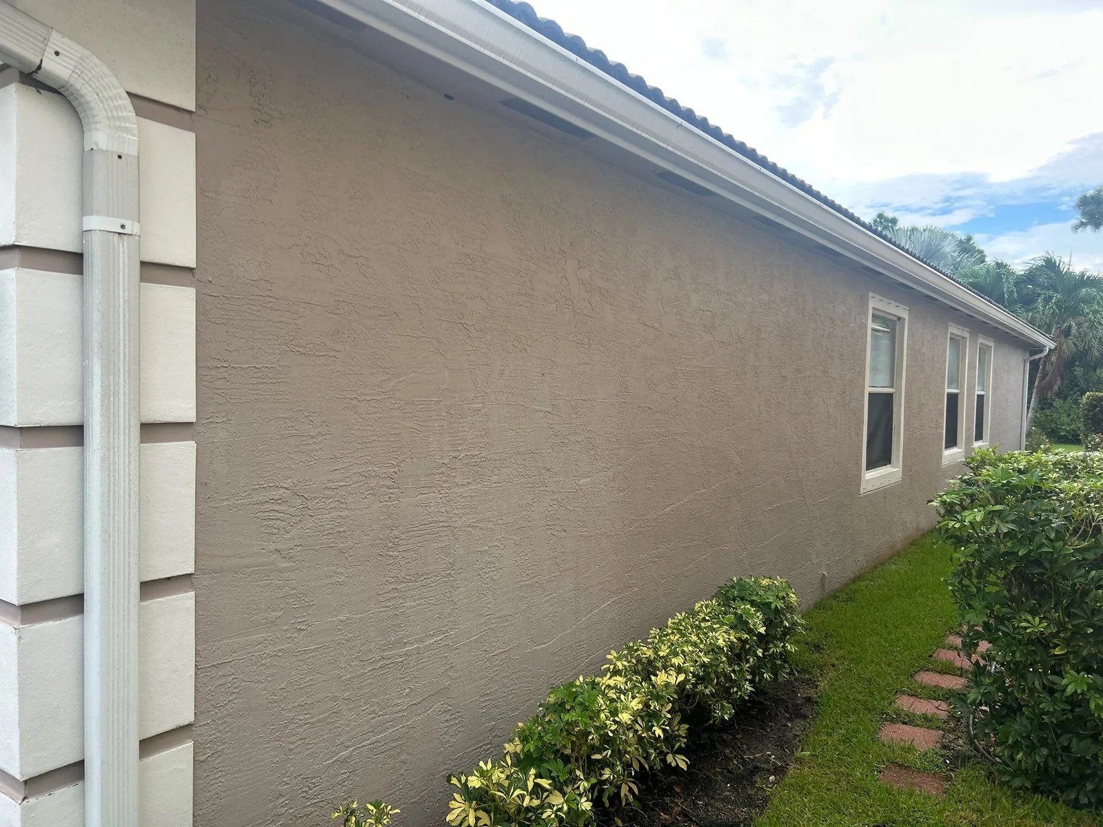Exterior Painting in Jensen Beach, Florida. Freshly painted tan stucco side elevation with white gutter and downspout, white-framed windows, hedging and lawn alongside.