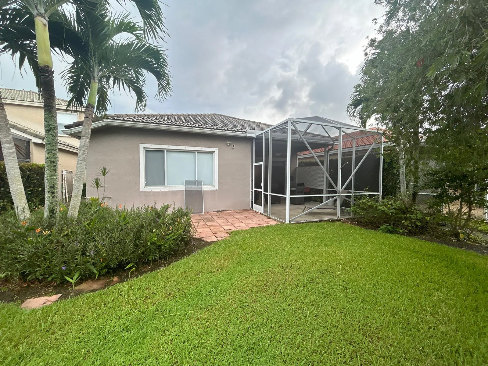 Exterior Painting in Jensen Beach, Florida. Rear elevation of a single-storey house with freshly painted grey-beige stucco, white trim, attached screen enclosure, paver patio and mown lawn.