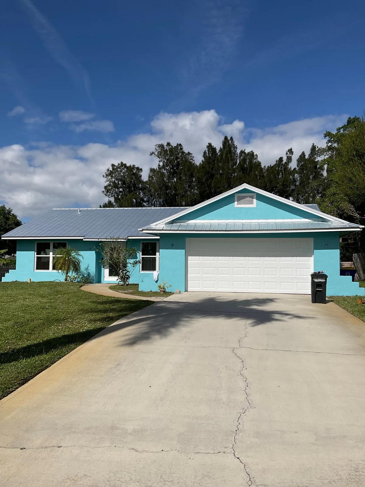 Exterior Painting in Jensen Beach, Florida. Single-storey house with turquoise painted exterior walls, white garage door and trim, and a metal roof, seen straight on from the driveway.