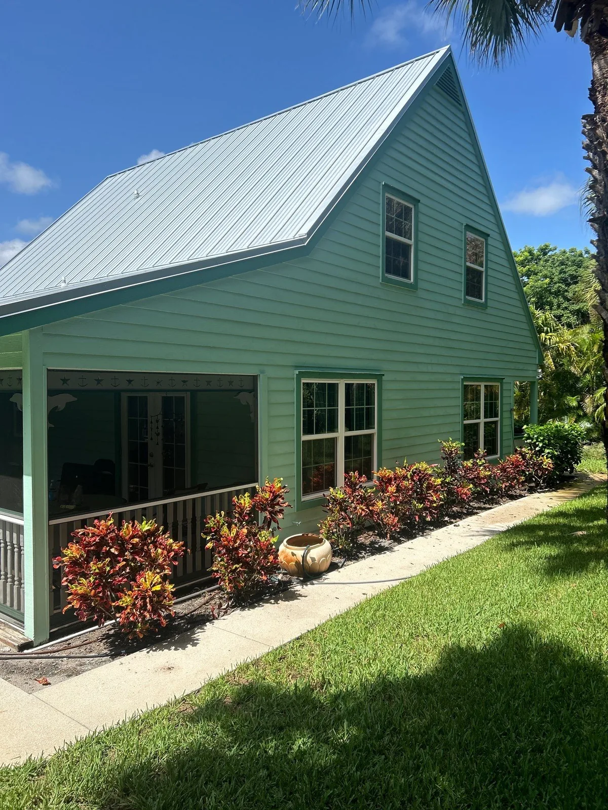 Exterior Painting in Jensen Beach, Florida. Two-storey green lap-siding house with white metal roof, white window trim and a screened porch, with croton plantings along a concrete walkway.