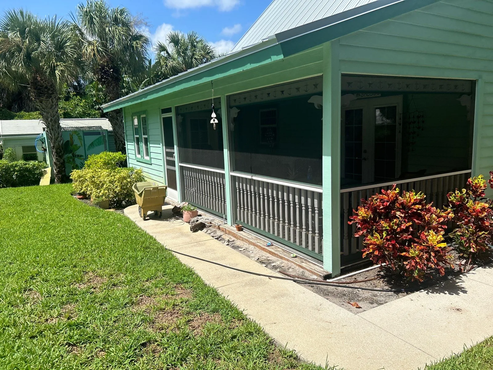 Exterior Painting in Jensen Beach, Florida. Angled view of a mint green house with a covered screened porch, dark railing and white posts, palms and croton plantings under a blue sky.