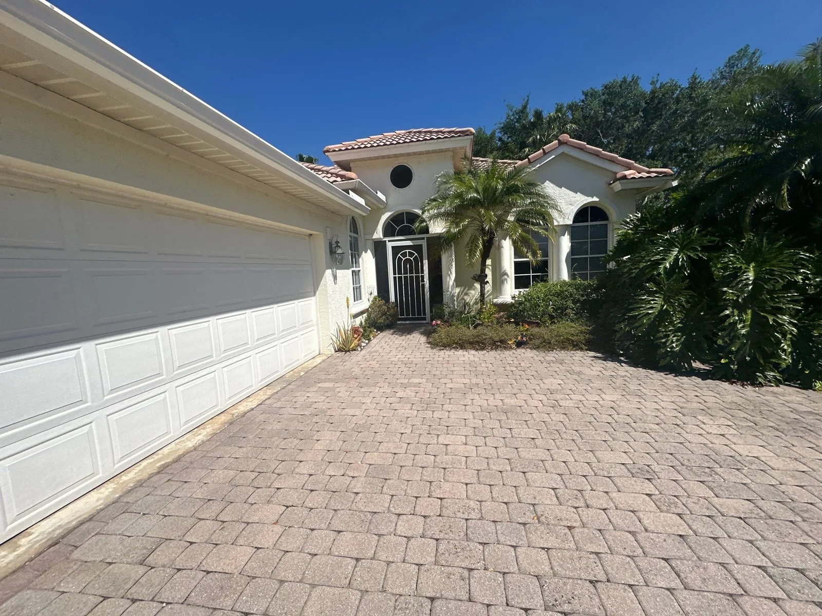 Exterior Painting in Jensen Beach, Florida. Single-storey home with barrel tile roof, painted light stucco exterior, white sectional garage doors and an arched entry, paver driveway in the foreground.