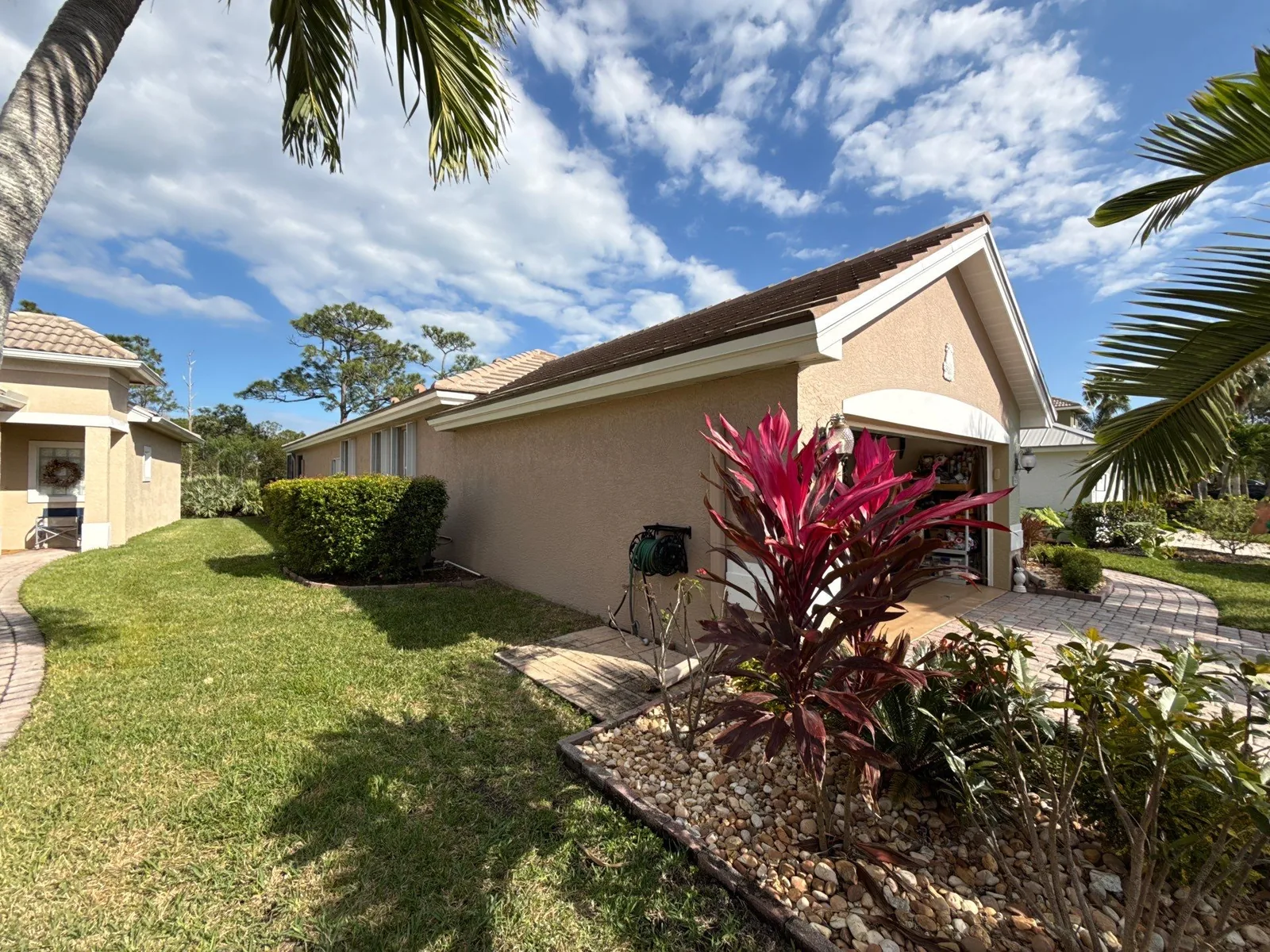 Exterior Painting in Jensen Beach, Florida. Single-storey home with tan stucco walls, darker trim and a tile roof, with lawn and landscaped bed in front.