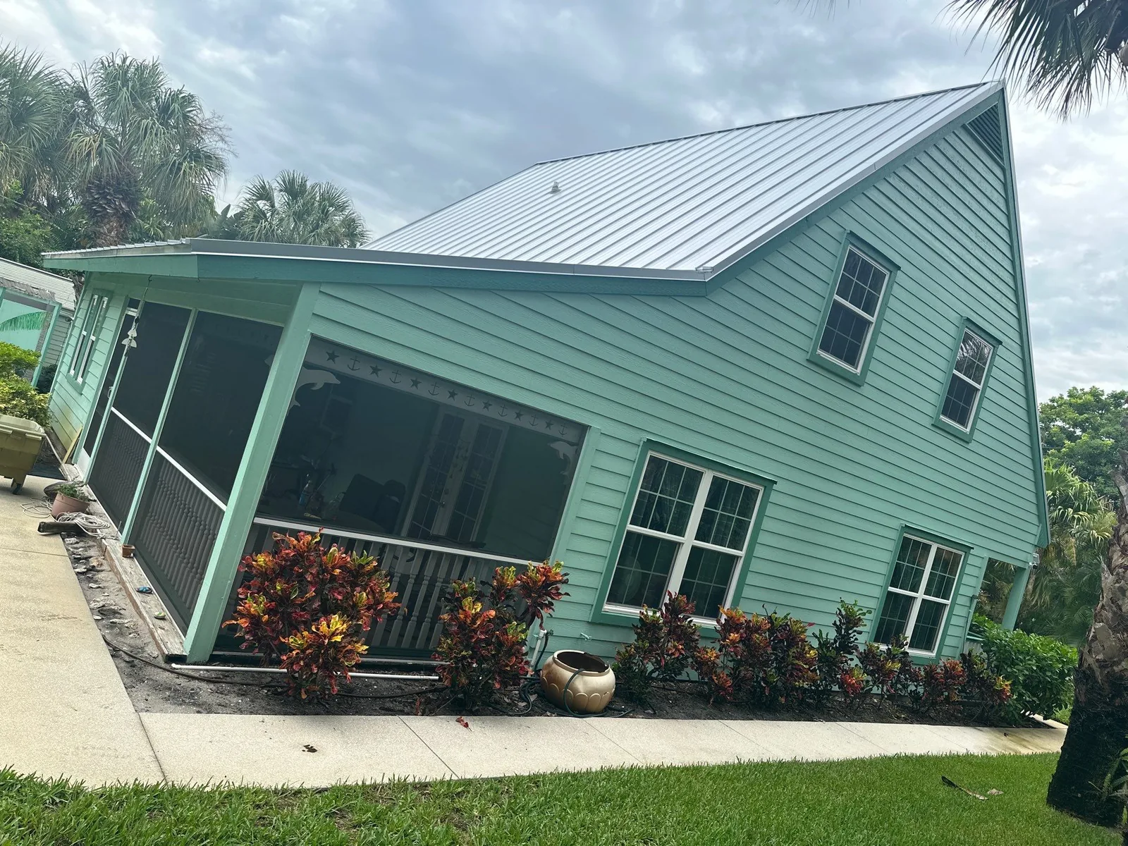 Exterior Painting in Jensen Beach, Florida. Corner of a house with pale green lap siding and white window trim, screened porch enclosure and standing-seam metal roof, croton shrubs along the wall.