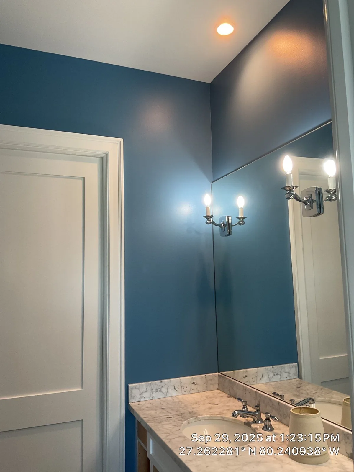 Interior Painting in Jensen Beach, Florida. Bathroom with deep blue walls, large frameless mirror, chrome wall sconces, white panelled door and stone-topped vanity.