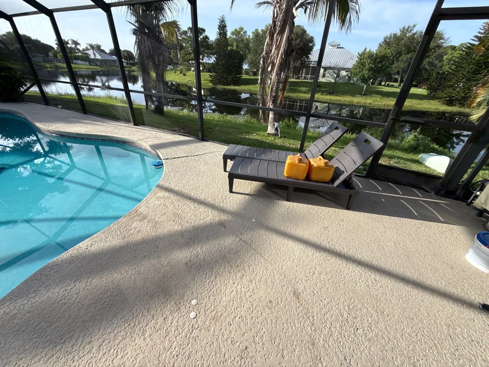 Pressure Washing in Jensen Beach, Florida. Clean screened pool deck with two loungers beside a kidney-shaped pool, overlooking a lawn and water.