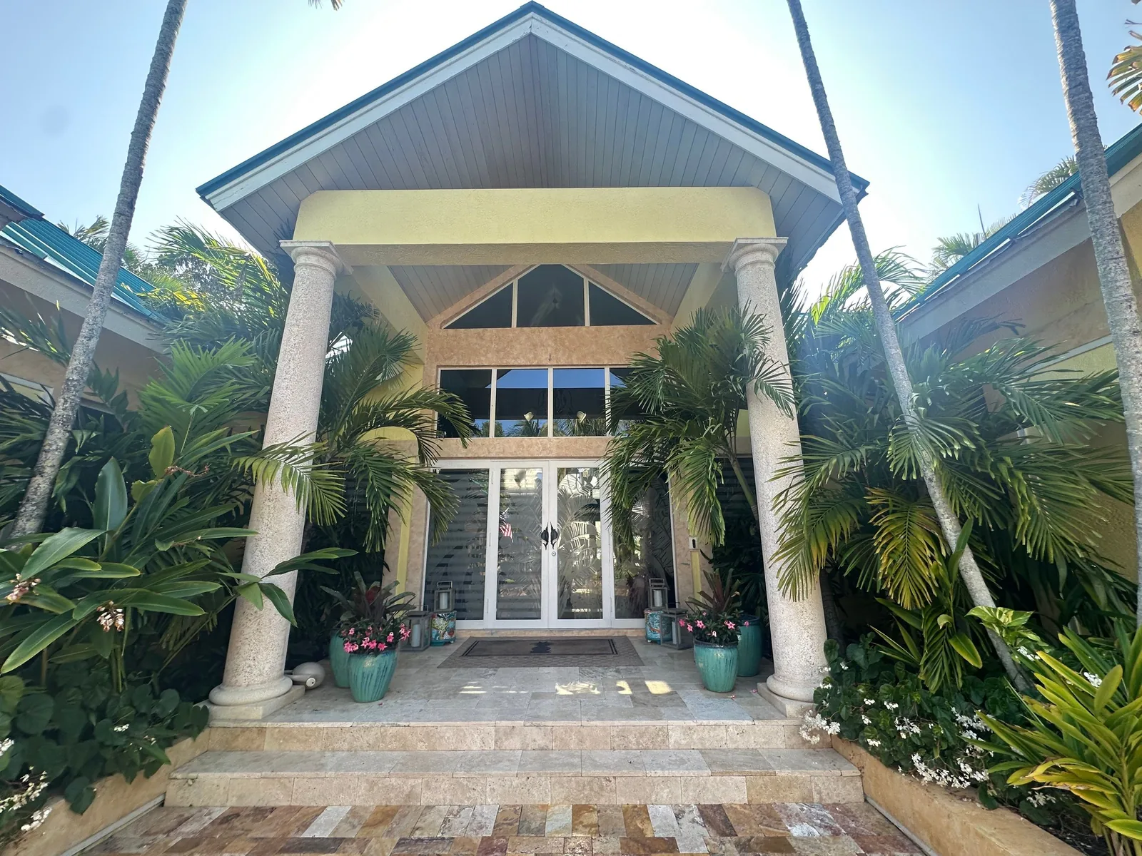 Exterior Painting in Juno Isles, Florida. Symmetrical front entrance of a home with cream columns, gabled portico, glass double doors and flanking palms under blue sky.