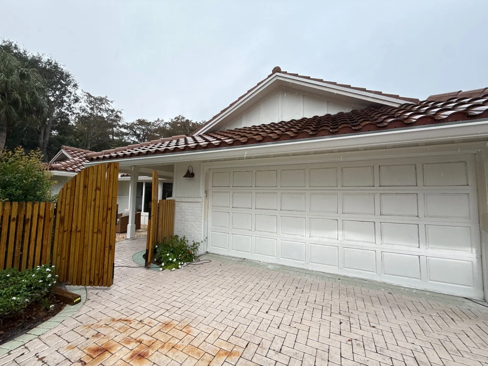 Exterior Painting in Juno Isles, Florida. Single-storey house with white painted exterior and trim, barrel tile roof, white sectional garage door, wood gate and paver driveway.