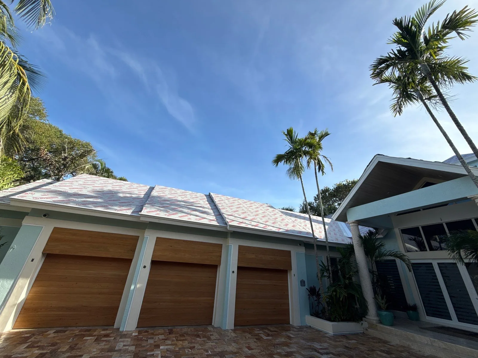 Exterior Painting in Juno Isles, Florida. Three-bay garage on a pale blue-green home with white trim and wood-finish garage doors, palm trees and a paver driveway.