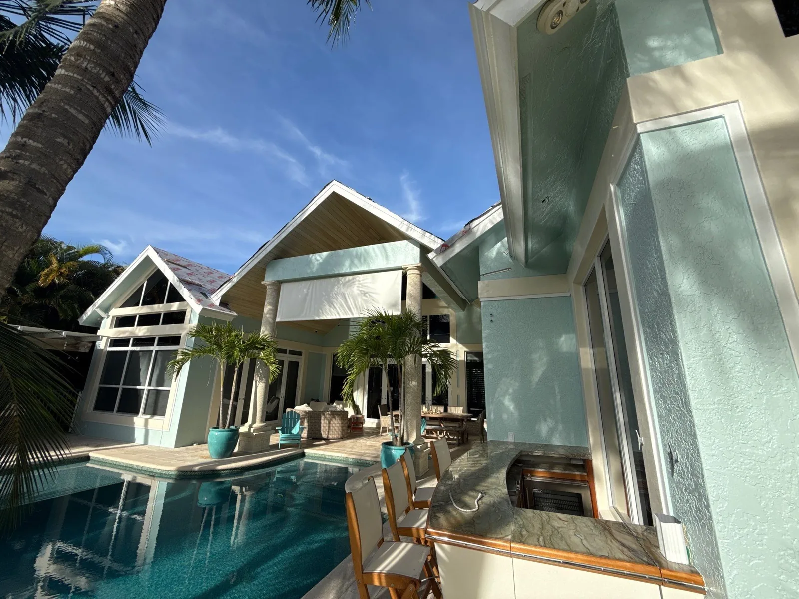 Exterior Painting in Juno Isles, Florida. Rear elevation of a pale blue-green home with white trim and gabled windows, with a swimming pool and patio in the foreground.