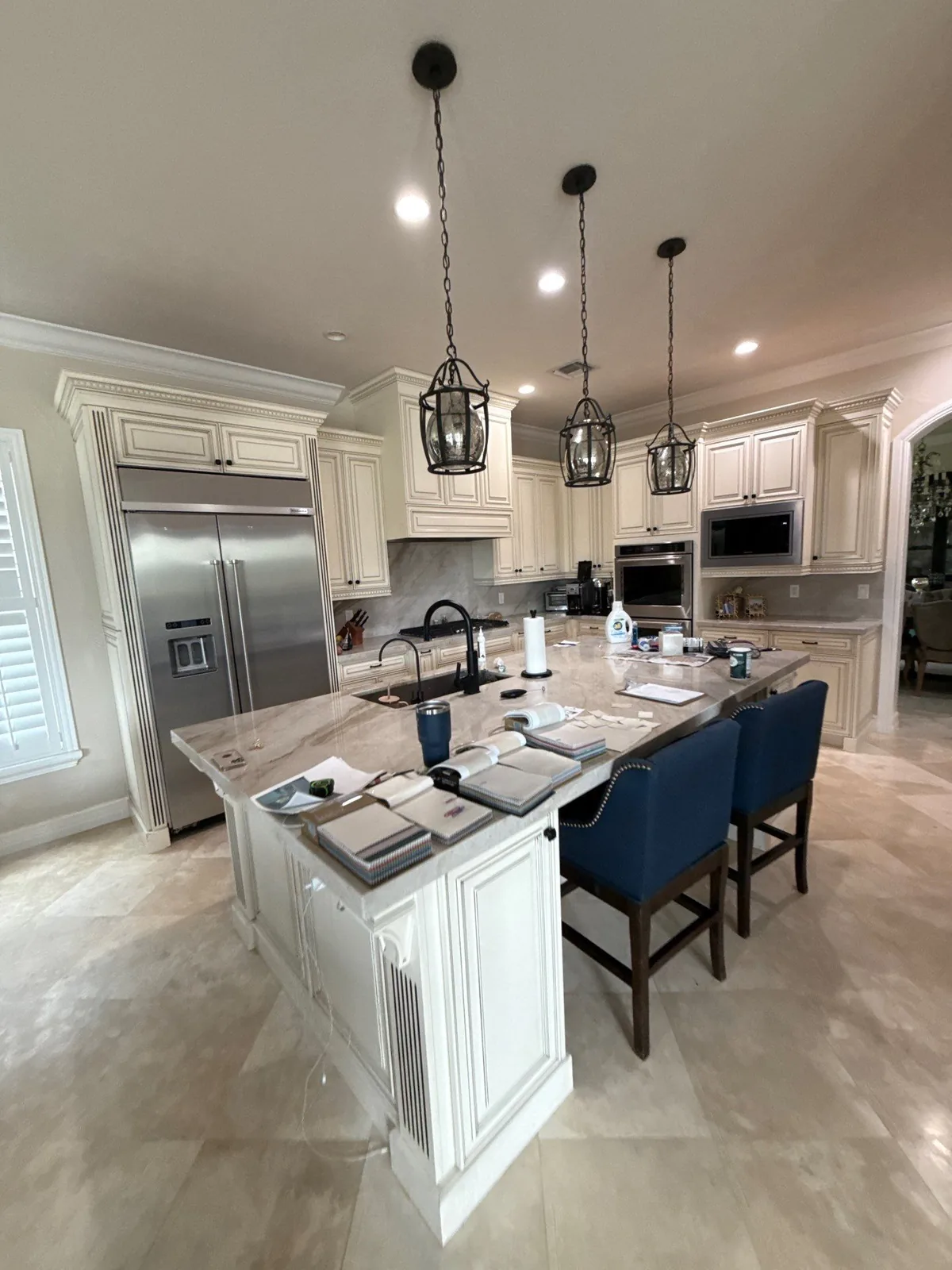 Cabinet Refinishing in Jupiter, Florida. Kitchen with cream glazed raised-panel cabinets, large stone-topped island, black lantern pendants and blue upholstered stools.