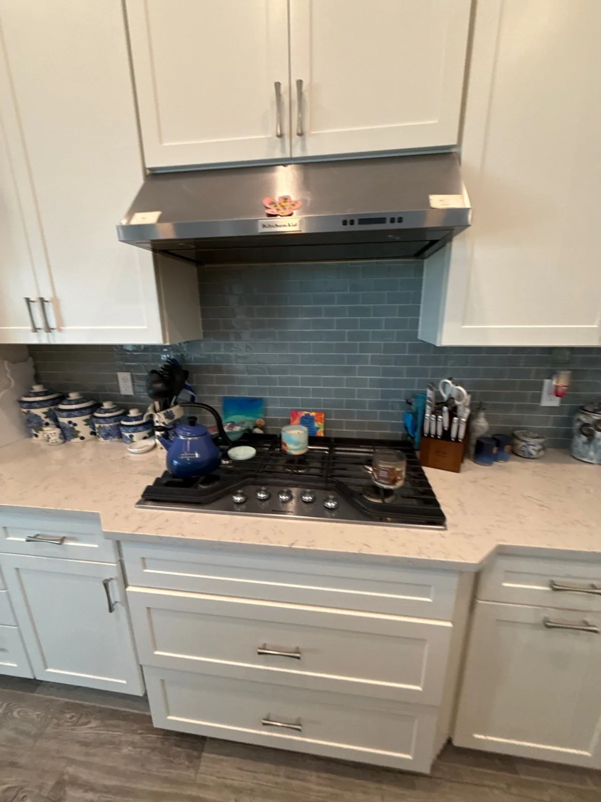 Cabinet Refinishing in Jupiter, Florida. White shaker kitchen cabinets around a stainless range hood and gas cooktop, with blue subway tile backsplash and light stone countertop.